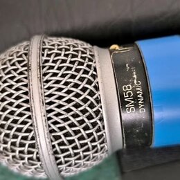 Detailní pohled na dynamický mikrofon s černým tělem a modrým kroužkem. Na stříbrném prstenci pod mřížkou je černý nápis SM58 DYNAMIC. Kovová mřížka…