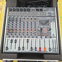 Stříbrný mixážní pult značky behringer, model XENYX X1222USB. Zařízení je umístěno v ochranném černém kufru s pěnovou výplní. Na panelu jsou vidět po…