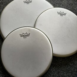 Tři bílé kruhové bubnové blány s kovovými obručemi. Každá blána má v horní části černé logo REMO a pod ním nápis EMPEROR® WHITE COATED MADE IN U.S.A.…