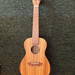 Dřevěný nástroj typu ukulele s přirozenou barvou a výraznou sletovanou strukturou dřeva. Na hlavě je viditelné logo ve tvaru stylizovaného písmena „C…
