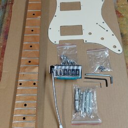Sada komponentů pro elektrickou kytaru položená na hnědé kartonové ploše. Obsahuje dřevěný krk s hnědým lakem, bílý plastový pickguard a bílou krytku…