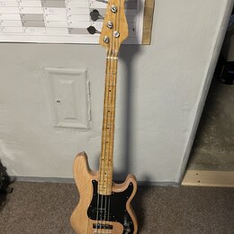 Elektrická basová kytara s přírodním dřevěným povrchem těla a hmatníku. Nástroj má černý pickguard, dva snímače a čtyři ladicí kolíky na hlavě. Na hl…