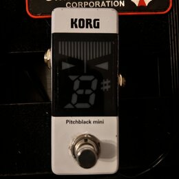 Bílý kytarový ladič s černým displejem. Na horní části je nápis KORG a pod displejem text Pitchblack mini. V horní části fotky je vidět nálepka s náp…