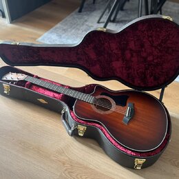 Akustická kytara s cutawayem v tmavě hnědém sunburst provedení, umístěná v otevřeném černém tvrdém pouzdře s červeným plyšovým vnitřkem. Na hlavě hma…