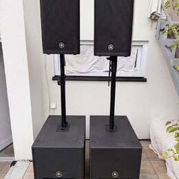 Dva černé reproduktory umístěné na černých stojaních, které jsou zapuštěny do dvou černých subwooferů. Všechny čtyři komponenty mají na přední straně…