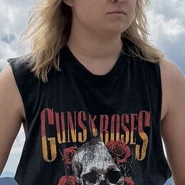 Fotografie zobrazuje osobu v černém tričku bez rukávů. Na přední straně trička je žlutý nápis GUNS N ROSES a grafika lebky s červenými růžemi. Osoba…