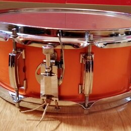 Pomalu vyfotocený oranžový snare drum s chromovaným obručováním a mechanismem. Na koruse nejsou viditelné žádné značky ani nápisy. Povrch nástroje vy…