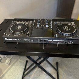 Černý DJ 컨트롤러 Numark MIXSTREAM PRO umístěný na černém stole. Na přední straně je vidět bílý nápis Numark MIXSTREAM PRO a menší text pod ním, který je…