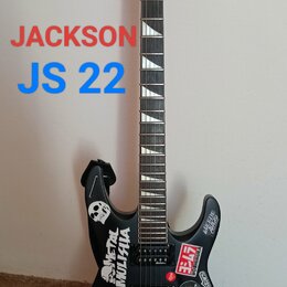 Černá elektrická kytara s hrotovitou hlavou, na které je bílé logo Jackson. Tělo kytary je polepeno různými nálepkami, včetně log Monster Energy a Ho…