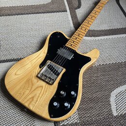 Elektrická kytara s přírodním dřevem a černým pickguardem. Na hlavě hnedku je viditelné logo Fender. Kytara má dva humbuckery, tři ovládací knofíky a…