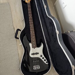 Černá elektrická basová kytara v otevřeném tvrdém kufru s černým vnitřkem. Na hlavě nástroje je viditelné logo Fender. Kytara má bílý pickguard, čtyř…