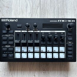 Černá groovebox s nápisy Roland a GROOVEBOX MC-101. Zařízení obsahuje displej, několik otočných ovladačů a bílá tlačítka s různými označením, včetně…