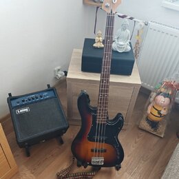 Elektrická basová kytara s tmavě hnědým sunburst povrchem a hmatníkem z tmavého dřeva. Na hlavě nástroje je bílý nápis Cort. Vedle kytary stojí malý…