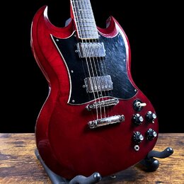 Elektrická kytara s červeným lakem a černým pickguardem, umístěná na černém stojanu. Nástroj má dva humbuckery a čtyři ovládací knoflíky. Povrch laku…
