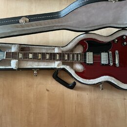 V otevřeném černém kufru s bílou vnitřní výplní leží červená elektrická kytara. Na hlavě nástroje je viditelné logo Gibson. Kytara má dva humbuckery,…
