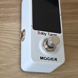 Bílý kytarový ladič v kompaktním kovovém provedení s černým displejem. Na přední straně jsou nápisy „Baby Tuner“ červenou barvou a pod spínacím tlačí…