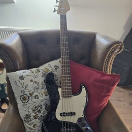 Černá elektrická basová kytara s bílým pickguardem a světlou hnedou hmatníkem. Na hlavě nástroje je viditelné logo Fender. Kytara je umístěna v hnědé…