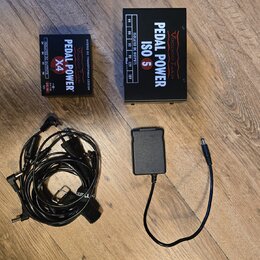 Na dřevěné podlaze jsou dvě černé napájecí zdroje pro efektové pedály a dva napájecí kabely. Menší zdroj má nápisy PEDAL POWER X4, ISOLATED DC OUTPUT…
