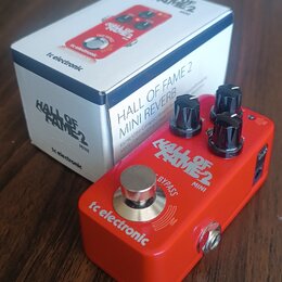 Červený efekt pedál s nápisy tc electronic, HALL OF FAME 2 MINI a BYPASS. Na horní straně jsou tři černé oválové ovládací knoflíky a jeden stříbrný k…