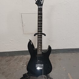 Černá elektrická kytara s špičatou hlavou, na které je bílé logo Jackson. Nástroj má tmavý hmatník a stříbrný kobylku typu Floyd Rose. Kytara je umís…