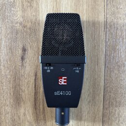 Černý dynamický mikrofon s kovovou mřížkou. Na těle jsou vidět přepínače s nápisy -10 0 -20 dB a 0 Hz, logo SE v červeném čtverci a modelové označení…