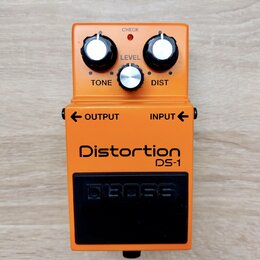 Oranžový kytarový efekt s nápisy Distortion a DS-1. Na horní části jsou tři ovládací knoflíky s popisky TONE, LEVEL a DIST oraz kontrolka CHECK. Ve s…