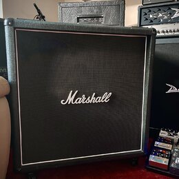 Černý kytarový reproduktor s bílým lemováním a logem Marshall uprostřed mřížky. V pozadí je vidět další černý reproduktor a kombo s nápisem Diezel. V…