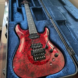Elektrická kytara s červeným mramorovaným povrchem těla v černém tvrdém kufru s modrou výstuhou. Nástroj má šest strun, dva humbuckery a dva ovládací…