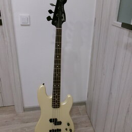 Elektrická basová kytara v krémové barvě s hnědým hmatníkem. Na hlavě nástroje je viditelné logo Fender. Tělo má dva snímače a čtyři ovládací knoflík…