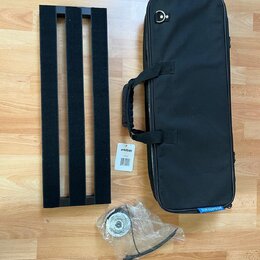 Na fotografii je černý kovový pedalboard s třemi vodicími lištami, černý přepravní vak s rukojetí a rolka kabelu v plastovém sáčku. Na vaku je modrý…