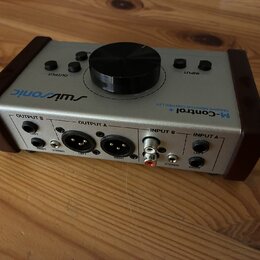Stříbrný monitor kontroler s hnědými bočními panely. Na horní straně je velký černý oválkový knoflík a nápisy „OUTPUT“, „INPUT“ a logo „swissonic“. N…