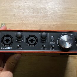 Červený audio rozhraní s černým předním panelem. Na zařízení jsou nápisy Scarlett 2i2, LINE INST, GAIN, DIRECT MONITOR, OFF ON, 48V a MONITOR. Jsou z…