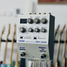 Bílý efekt pedál s nápisy BOSS, DIGITAL DELAY a DD-200. Zařízení má pět ovládacích knoflíků s popisky TIME, FEEDBACK, E. LEVEL, PARAM, TONE a MOD DEP…