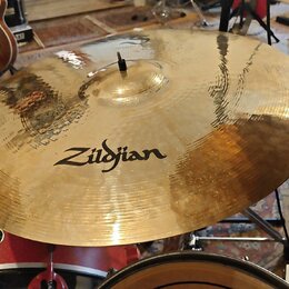 Zlatavý kovový činel s viditelným kovánívým povrchem a mírným opotřebením. Uprostřed je černý nápis Zildjian. Činel je osazen na stojanu v rámci bicí…