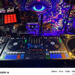 Fotografie zachycuje DJ set umístěný před barevným gobelínem s motivem oka. Uprostřed je černý DJ kontrolér se značkami RANE a serato, vybavený dvěma…
