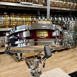 Vínový malý buben s chromovaným obručováním a mechanismy, umístěný na chromovaném stojanu. Na korpusu je zlatý štítek s nápisy TAMA, SNARE DRUM a Ser…