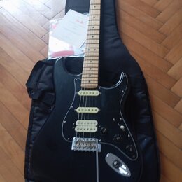 Černá elektrická kytara typu Stratocaster s bílým pickguardem a světlou hnedou hmatníkem. Na hlavě kytary je viditelné logo Fender a v pozadí leží pa…