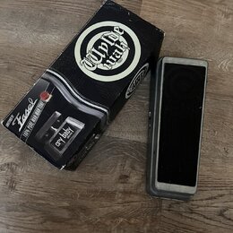 Na fotografii je wah-wah pedál v šedé barvě s černou gumovou plochou a vedle něj originální černá krabice. Na krabici je bílé logo „Dunlop“ a nápisy…
