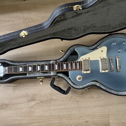 Modrá elektrická kytara v černém tvrdém kufru. Na hlavě je viditelné logo Epiphone. Kytara má dva humbuckery, bílý pickguard a čtyři ovládací knoflík…
