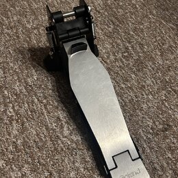 Kovový pedál s černým plastovým mechanismem, položený na hnědém koberci. Na spodní části stříbrného šlapáku je vyryté logo Roland. Povrch kovové část…