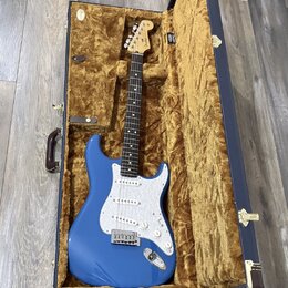 Modrá elektrická kytara typu Stratocaster s bílým pickguardem, uložená v otevřeném tmavomodrém kufru s hnědým plyšovým vnitřkem. Na hlavě kytary je v…