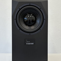 Černý studiový subwoofer v hranatém korpusu s matným povrchem. Na přední straně je umístěn velký kruhový reproduktor a pod ním ovládací panel s nápis…