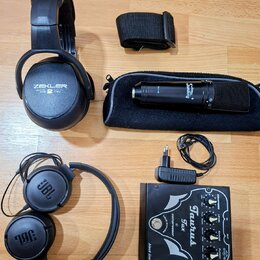 Na fotografii je sada audio vybavení: dvě černá sluchátka, mikrofon s pouzdrem a efektový pedál. První sluchátka mají nápis ZEKLER, druhá nesou logo…