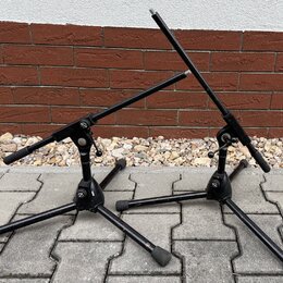Dvě černé kovové stojany na noty nebo mikrofony s trojnožkou a gumovými koncovkami. Na kloubech obou stojanů je viditelné malé kulaté logo s nápisem…