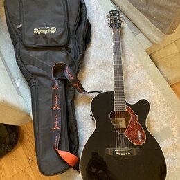 Černá akustická kytara s vyřezem v těle a hnědým rýhovaným plektrem. Na hlavě nástroje je nápis GRETSCH a na plektru jsou dvě loga GRETSCH. Kytara je…