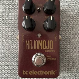 Bordeaux červený efekt pedál s nápisy MOJOMOJO a OVERDRIVE. Na spodní části je logo tc electronic a text true bypass. Pedál má čtyři černé ovládací k…