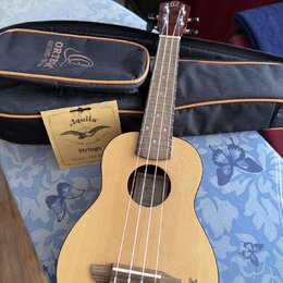 Světlý dřevěný ukulele s tmavým hmatníkem a čtyři struny. Na hlavě nástroje je viditelné logo ve tvaru stylizovaného písmene O. Součástí je černý pou…