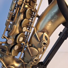Detail saxofonu v matné zlaté barvě, který je umístěn na černém stojanu. Na korpu nástroje je vyryto logo SELMER PARIS a nápis MADE IN FRANCE. Pod ní…