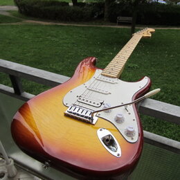 Elektrická kytara s tělem v barvě sunburst a bílým pickguardem. Na hlavě hředu je viditelné logo Fender. Nástroj má tři snímače, tři ovládací knoflík…