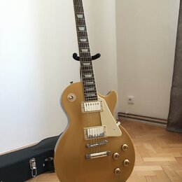 Zlatá elektrická kytara s tvarem singlecut, bílým pickguardem a dvěma humbuckery. Na hlavě hředu je viditelné logo Epiphone. Nástroj je umístěn na če…